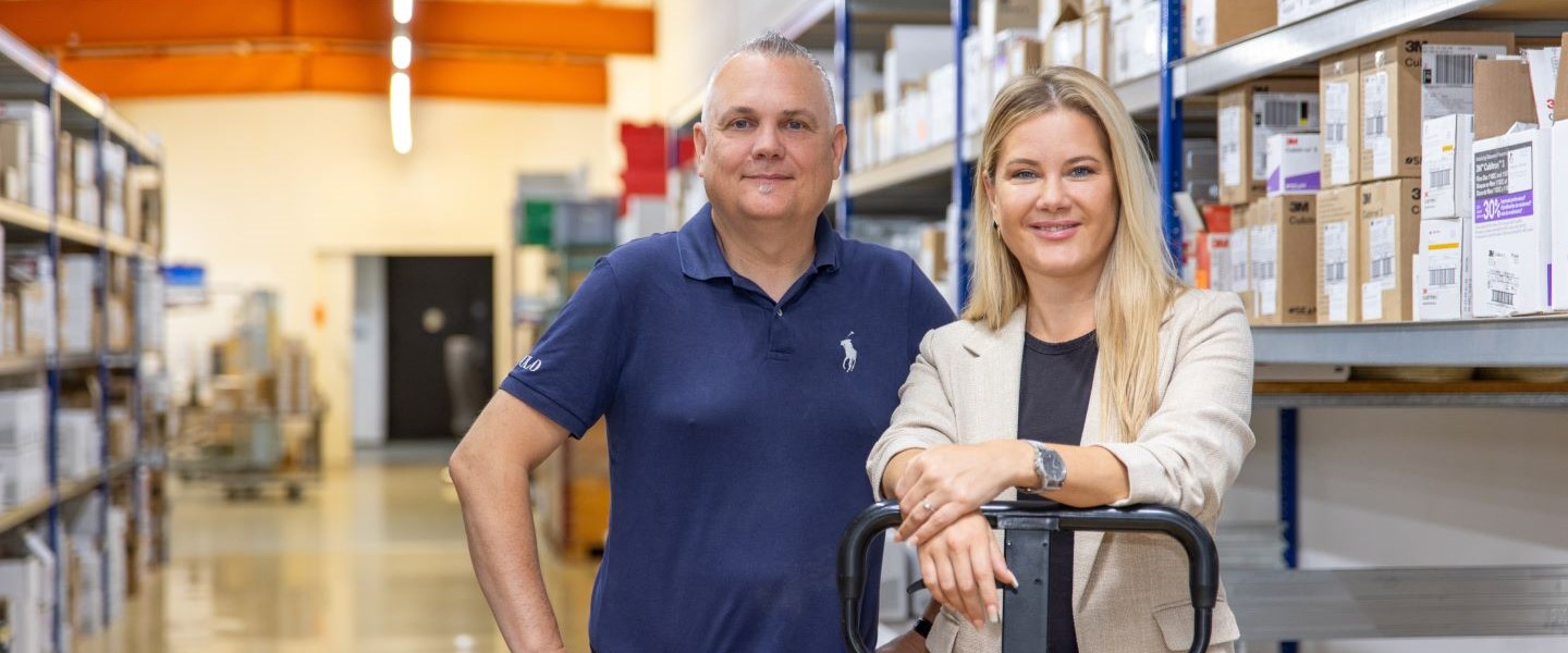 Lagerarbeiter Verkauf Wir sehen zwei Personen die in die Kamera schauen und in einer Lagerhalle stehen