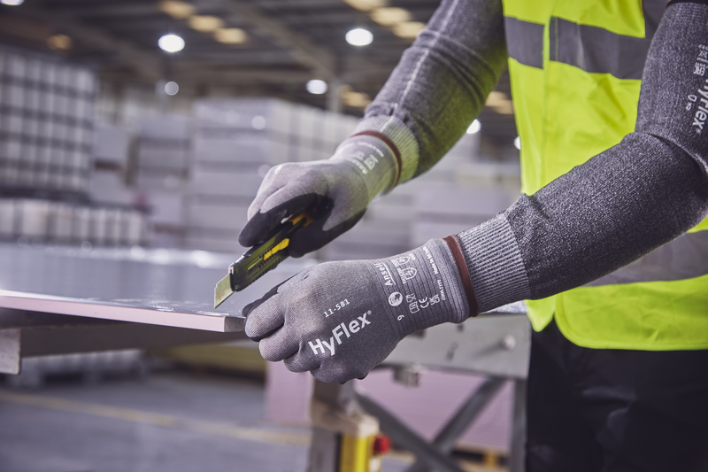 Gros plan sur une personne portant des gants anti-coupures HyFlex gris et un gilet de sécurité jaune, découpant avec précision une plaque claire au cutter. Mains protégées, lame suivant le bord. Stockage en arrière-plan.