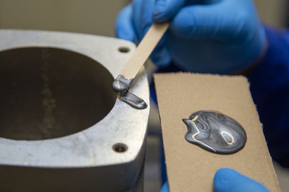 Eine Person mit blauen Handschuhen trägt silberfarbenen, pastösen Aluminium-Minutenkleber mit einem Holzspatel auf die Kante eines zylindrischen Aluminiumteils auf. Rechts liegt gemischter Klebstoff auf Karton, bereit zur Spaltfüllung.