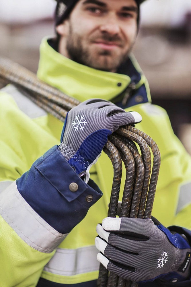 Ein Arbeiter in Warnkleidung und wasserdichten Tegera-Kälteschutzhandschuhen hält Bewehrungsstahl. Die grauen Handschuhe mit blauen Akzenten und weißen Fingerspitzen zeigen ein Schneeflockensymbol für Kälte und Nässe.