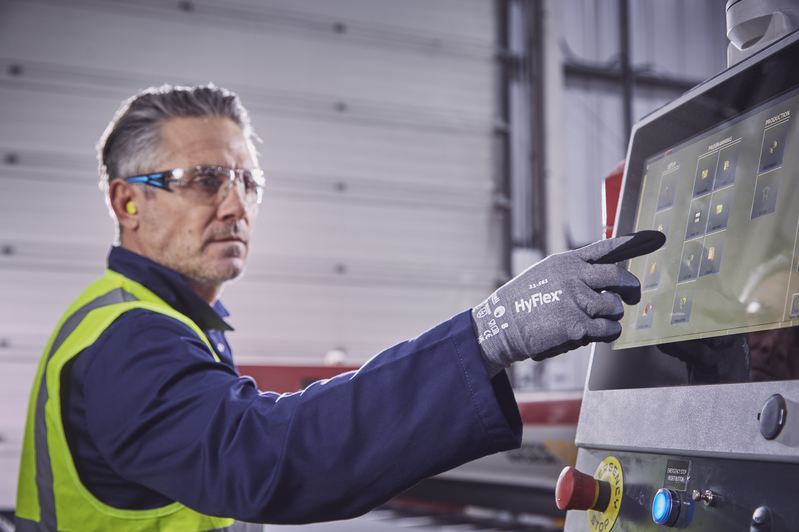 Dans un atelier, un ouvrier avec lunettes, casque antibruit et gilet de sécurité utilise l'écran tactile d'une machine. Il porte des gants HyFlex gris anticoupures compatibles tactile et sélectionne une option.