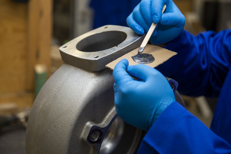 Person mit blauen Handschuhen rührt auf Karton mit Holzspatel eine metallisch-graue Paste an: WEICON Alu-Minutenkleber, aluminiumgefüllter Epoxidharzklebstoff zur Verklebung/Reparatur eines Aluminiumteils im Hintergrund.