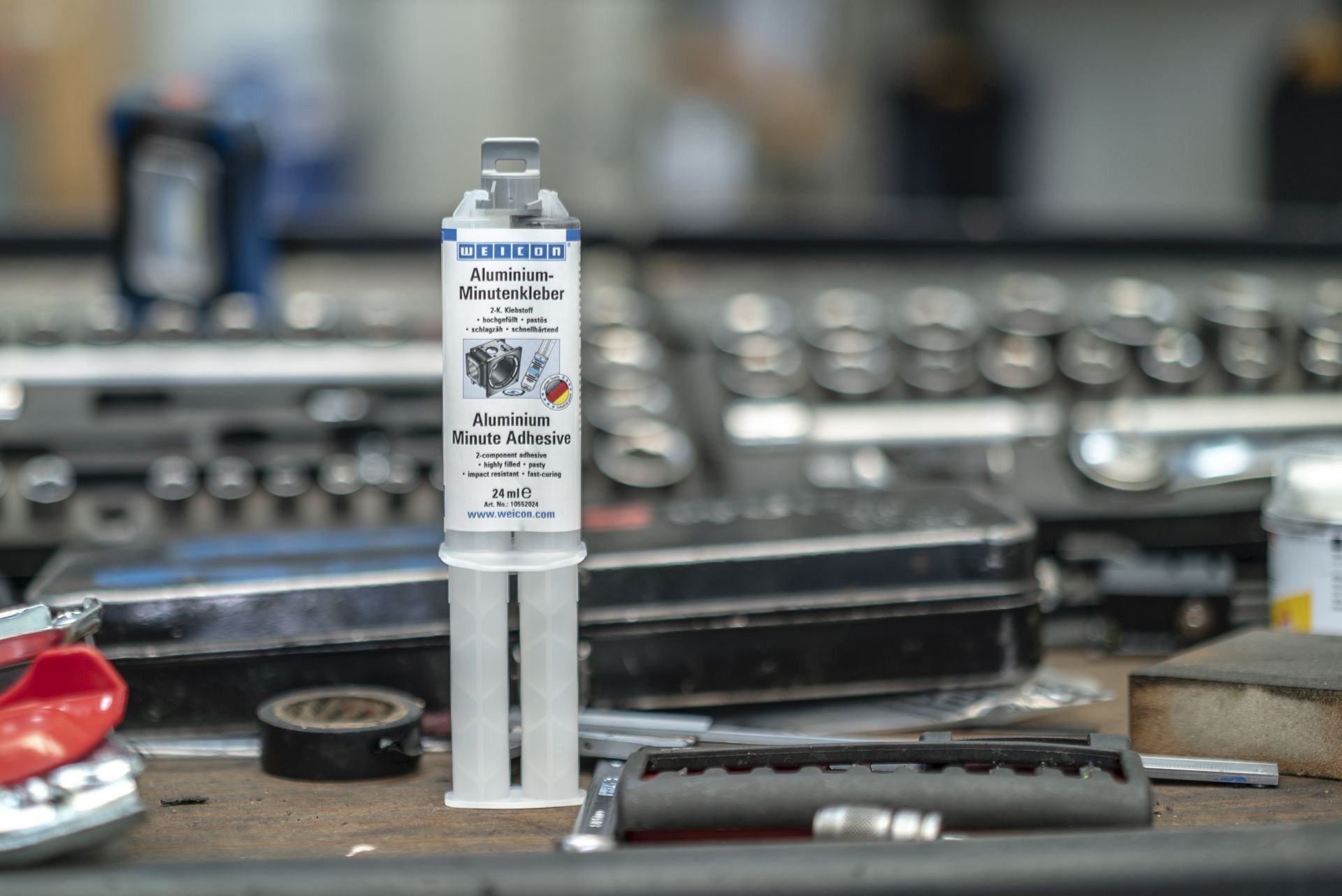 WEICON Aluminium-Minutenkleber in aufrechter Doppelkammer-Tube steht auf einer hölzernen Werkbank. Das Etikett beschreibt den pastösen, schnellhärtenden, aluminiumgefüllten 2K-Klebstoff als spaltfüllend für Alu.